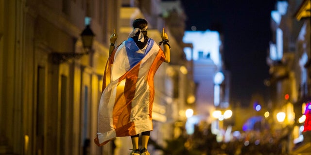 In this Wednesday photo, a demonstrator with a Puerto Rican flag draped over his shoulders balances on the hands of another during clashes in San Juan, Puerto Rico. (AP Photo/Dennis M. Rivera Pichardo)