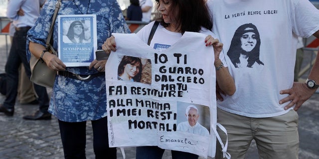 Reanta Grattani, who claims to be friends with the Orlandi family, middle, holds a T-shirt with the pictures of Emanuela Orlandi and Pope Francis and a written reading, "I'm watching you from heaven, but you (referring on Pope Francis), please let her return my mortal remains to my mother. " outside the Vatican, Saturday. (AP Photo / Gregorio Borgia)