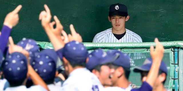 In this July 25, 2019, photo, Ofunato High School pitcher Roki Sasaki on the bench watches Hanamaki Higashi High School players celebrate their 12-2 win in their final baseball game at the Iwate Prefecture qualifier in Morioka, northern Japan. (Kyodo News via AP)