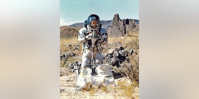 Gordon Swann in suit holding Apollo Lunar Staff prototype; tool carrier in front