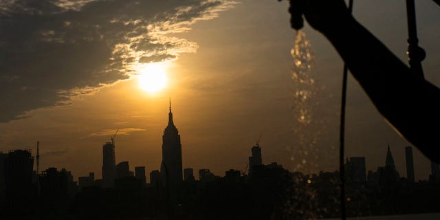 The sun rises over New York City and the Empire State Building while a man sprays water at Pier A on Saturday, July 20, 2019 in Hoboken, N.J.