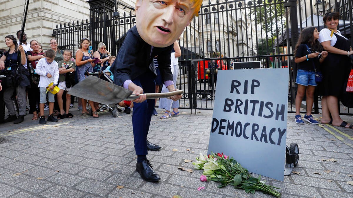 A man in a giant Boris Johnson "head" digs a grave at the foot of a pretend tombstone outside Downing Street in London, Wednesday, Aug. 28, 2019. 