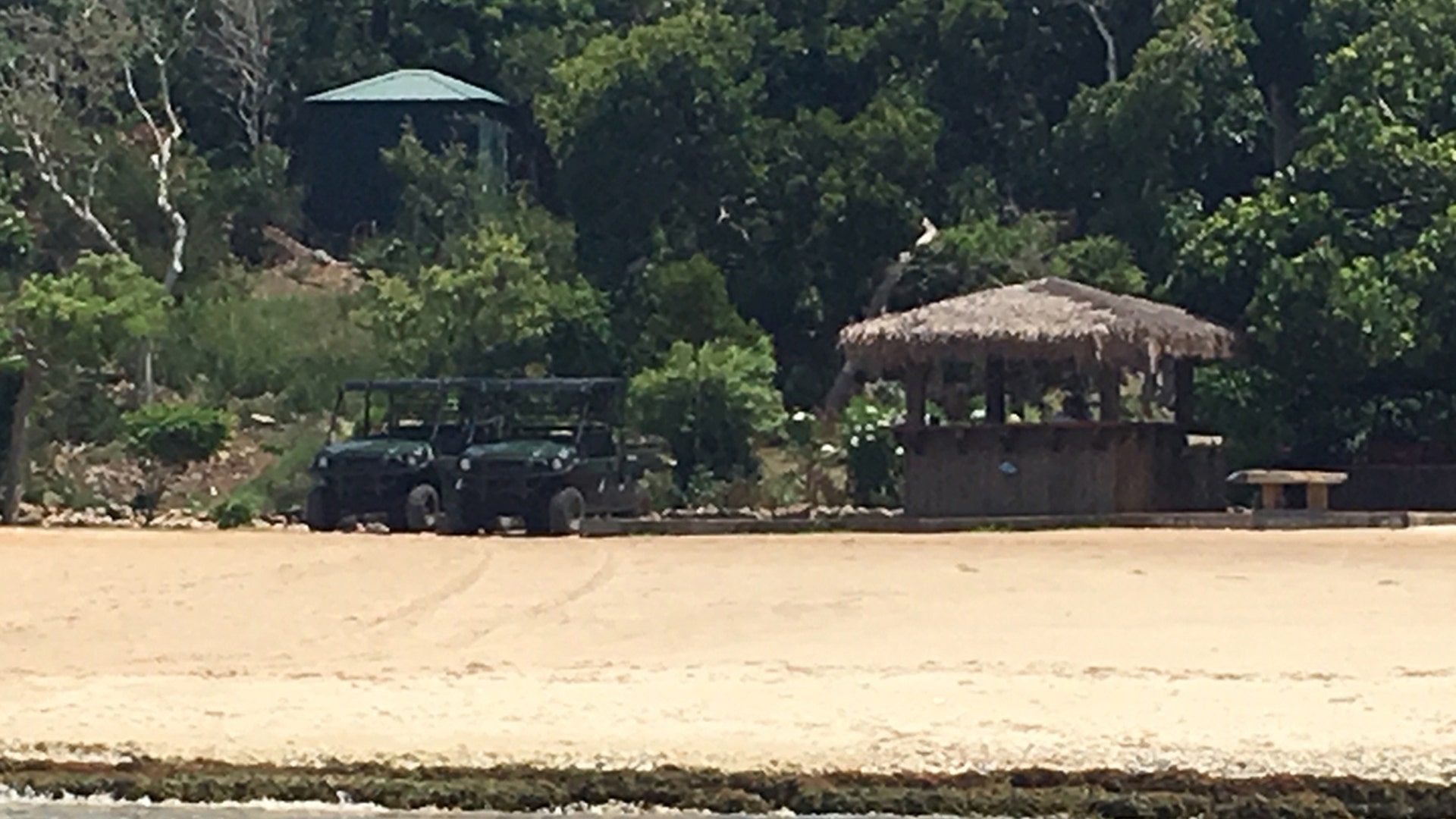 Two hunter green empty construction carts are next to a staw roof structure. There is a lone security guard in a white shirt sitting inside. 