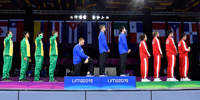In this Friday, Aug. 9, 2019 photo, released by Lima 2019 News Services, Race Imboden of the United States takes a knee, as teammates Mick Itkin and Gerek Meinhardt stand on the podium after winning the gold medal in team's foil, at the Pan American Games in Lima, Peru. (Jose Sotomayor/Lima 2019 News Services via AP)