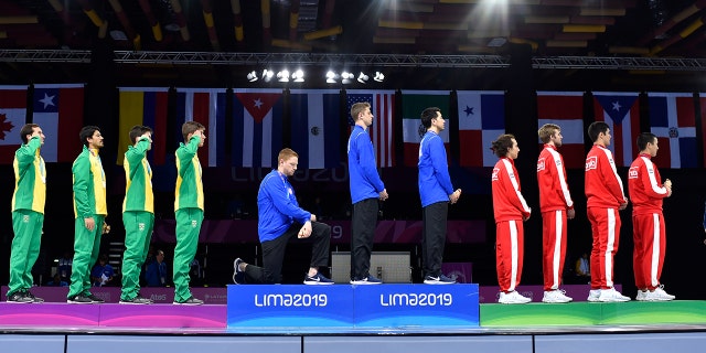 In this Friday, Aug. 9, 2019 photo, released by Lima 2019 News Services, Race Imboden of the United States takes a knee, as teammates Mick Itkin and Gerek Meinhardt stand on the podium after winning the gold medal in team's foil, at the Pan American Games in Lima, Peru. (Jose Sotomayor/Lima 2019 News Services via AP)