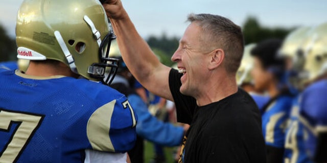 Coach Joe Kennedy kneels on the football field joined by his players.