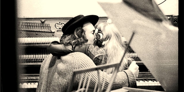 David Crosby holding Joni Mitchell while both are seated at an upright piano, seen from the back, through the control room window at the recording studio. — Getty