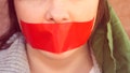 Young woman with serious expression is looking at the camera. She is wearing a piece of red tape over her mouth as a sign of silent protest. Activist is wearing a green head scarf. A second young woman is also wearing tape over her mouth in the background, while leaning against a stone wall.