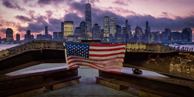 A U.S. flag hanging from a steel girder, damaged in the Sept. 11, 2001 attacks on the World Trade Center, blows in the breeze at a memorial in Jersey City, N.J., Sept. 11, 2019 as the sun rises behind One World Trade Center building and the re-developed area where the Twin Towers of World Trade Center once stood in New York City on the 18th anniversary of the attacks. (AP Photo/J. David Ake)
