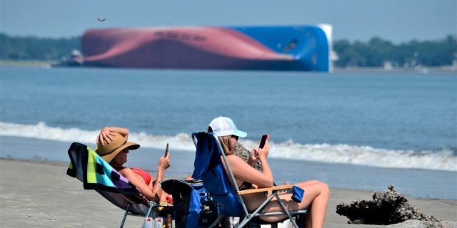 Rachel, left, and Sarah Mitchell look at their phones as they sunbathe on Jekyll Island's Driftwood Beach as the Golden Ray cargo ship is capsized in the background, off the Georgia coast, Sunday, Sept. 8, 2019.