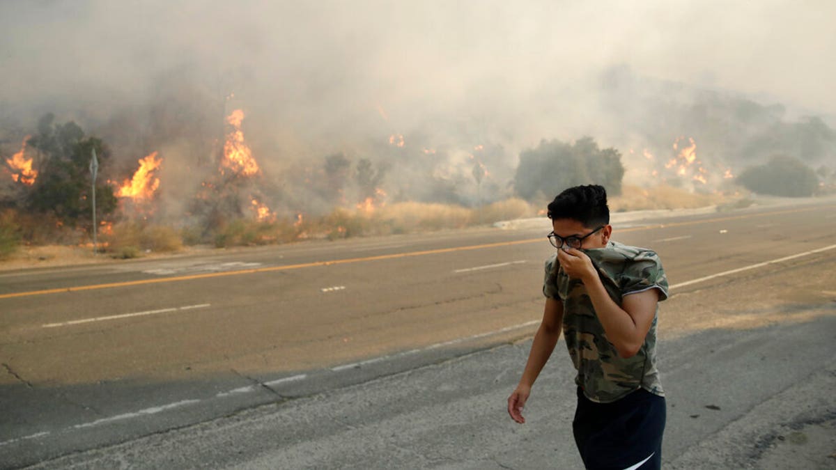 Brandon Mani covers his face from the smoke created by a wildfire as he walks along Highway 14 Thursday, Oct. 24, 2019, in Santa Clarita, Calif. (AP Photo/Marcio Jose Sanchez)