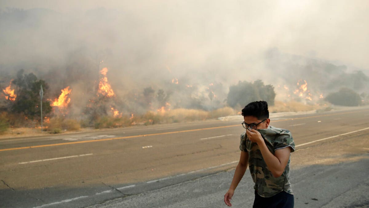 Brandon Mani covers his face from the smoke created by a wildfire as he walks along Highway 14 Thursday, Oct. 24, 2019, in Santa Clarita, Calif. (AP Photo/Marcio Jose Sanchez)
