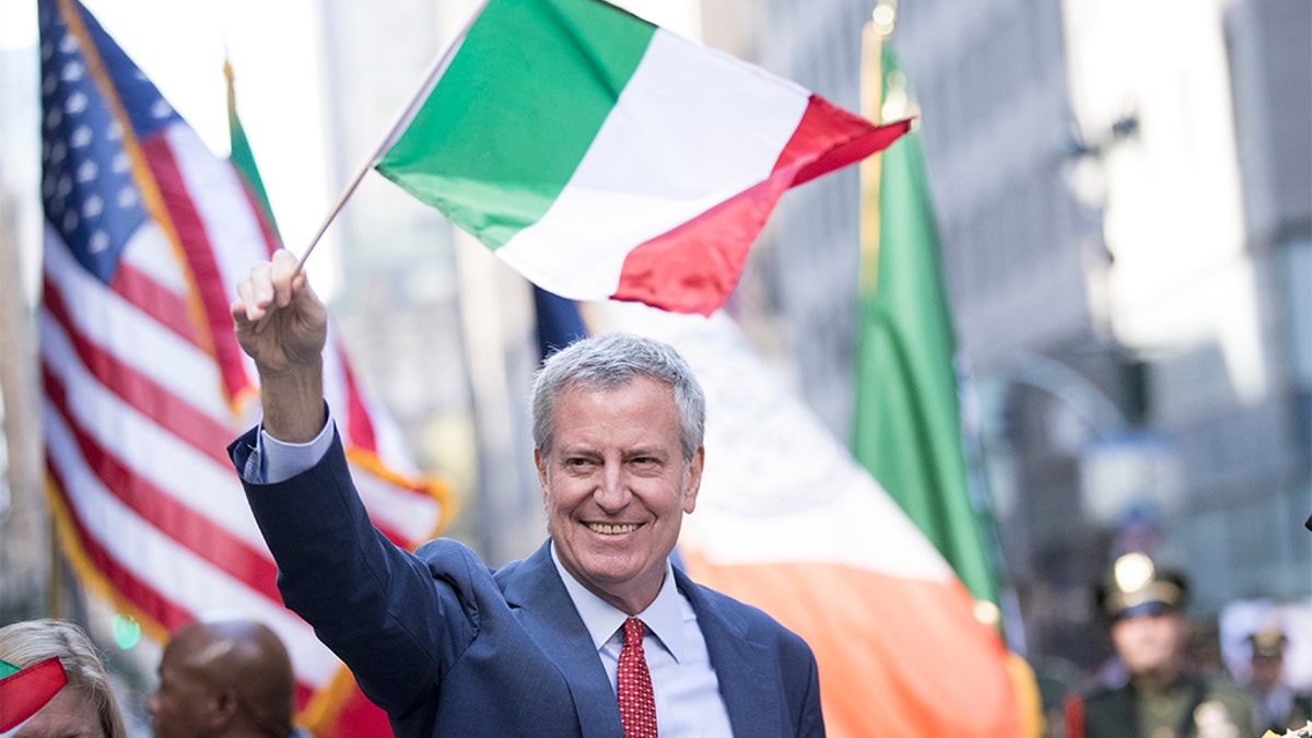 New York City Mayor Bill de Blasio waves to people along the street while holding an Italian flag. (Photo by Ira L. Black/Corbis via Getty Images)