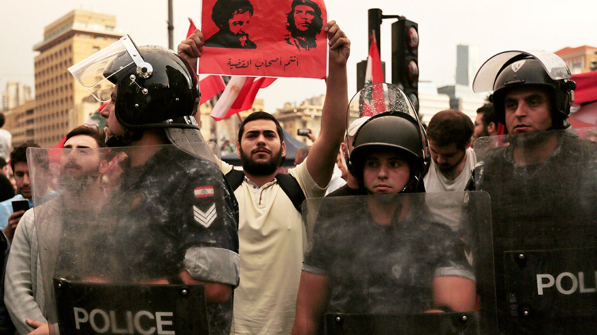 An anti-government protester holds up a placard with the pictures of Shiite cleric Imam Moussa al-Sadr, left, who went missing with his two companions and Che Guevara with Arabic that reads "You are the cause owner," as Lebanese riot policemen separates between anti-government protesters, background, and Hezbollah supporters during a protest in Beirut, Lebanon. (AP Photo/Hassan Ammar)
