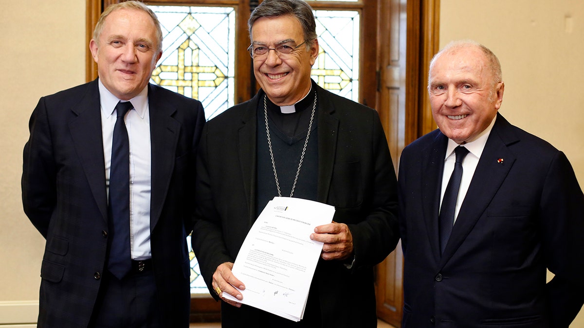 Francois-Henri Pinault, left, his father Francois Pinault, right, and Archbishop of Paris Michel Aupetit, center, pose after signing an agreement to raise money for the rebuilding of Notre-Dame cathedral. (AP Photo/Thibault Camus)