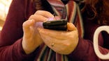 A close up, table-top view of the hands of a Caucasian woman, taken in front of her as she uses a smartphone during a coffee break - a blue LCD light glows in her hands. The woman wears a woolly scarf &amp; jumper and part of a mug is seen in the frame.