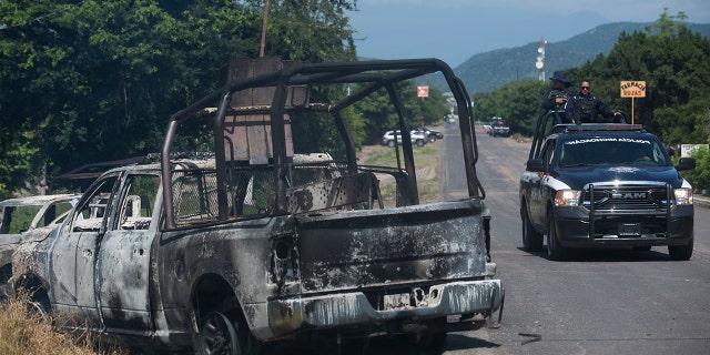 A charred truck that belongs to Michoacan state police sits on the side of the road after it was burned during an attack, as state police drive past in El Aguaje, Mexico, Monday, Oct. 14, 2019. At least 13 police officers were killed and three others injured Monday in the ambush. (AP Photo/Armando Solis)