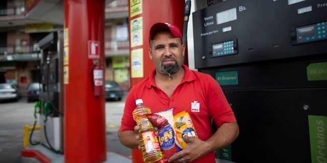 In this Oct. 8, 2019 photo, gas station attendant Leowaldo Sanchez poses with food items he was paid with by motorists: a bottle of cooking oil, a kilogram of rice and a package of corn flour, as he works at the pump in San Antonio de los Altos on the outskirts of Caracas, Venezuela. Bartering at the pump has taken off as hyperinflation makes Venezuela’s paper currency, the bolivar, hard to find and renders some denominations all but worthless, so that nobody will accept them. (AP Photo/Ariana Cubillos)