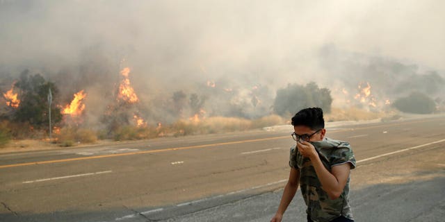 Brandon Mani covers his face from the smoke created by a wildfire as he walks along Highway 14 Thursday, Oct. 24, 2019, in Santa Clarita, Calif. (AP Photo/Marcio Jose Sanchez)