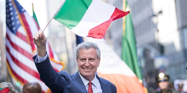 New York City Mayor Bill de Blasio waves to people along the street while holding an Italian flag. (Photo by Ira L. Black/Corbis via Getty Images)