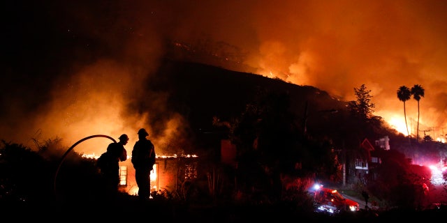 In this Dec. 7, 2017, file photo, two firefighters watch as a home burns in a wildfire in La Conchita, Calif.