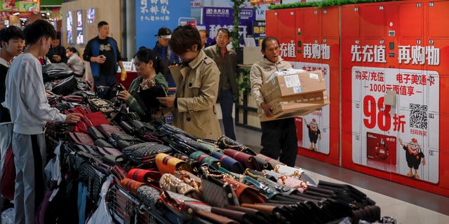 A worker carrying boxes of goods walks by women selecting handbags at a commercial building in Beijing, Friday, Oct. 18, 2019. (AP Photo/Andy Wong)