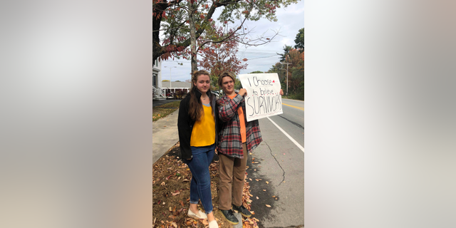 In this Monday, Oct. 7, 2019 photo provided by Shael Norris, high school sophomore Aela Mansmann, 15, of Cape Elizabeth, Maine, left, stands with her brother Aidan, 13, as he displays a placard during a school walkout, in Cape Elizabeth. The American Civil Liberties Union of Maine is calling on a federal court to stop the suspension of Aela Mansmann who accused an unnamed person of sexual assault. Aela and Aidan participated in the school walkout meant to protest the suspension of Aela and two other students. (Shael Norris via AP)