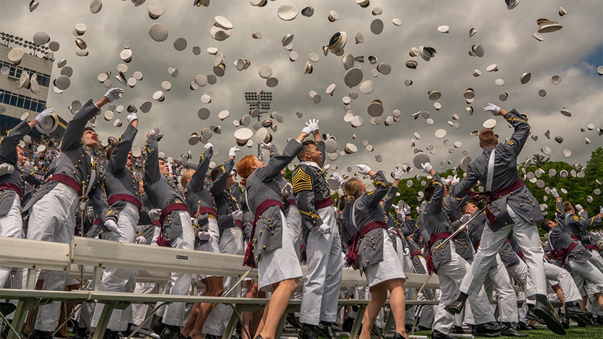 West Point graduates in gray uniforms celebrate commissioning by throwing hats in the air