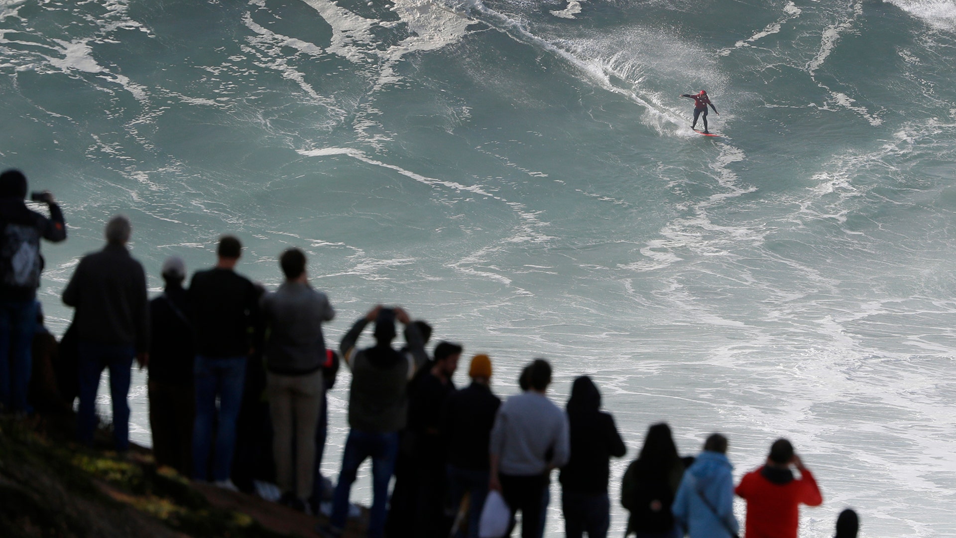 People on top of a cliff watch as a surfer rides a wave during a big wave surfing session at the Praia do Norte, or North Beach, in Nazare, Portugal, Nov. 20, 2019. 