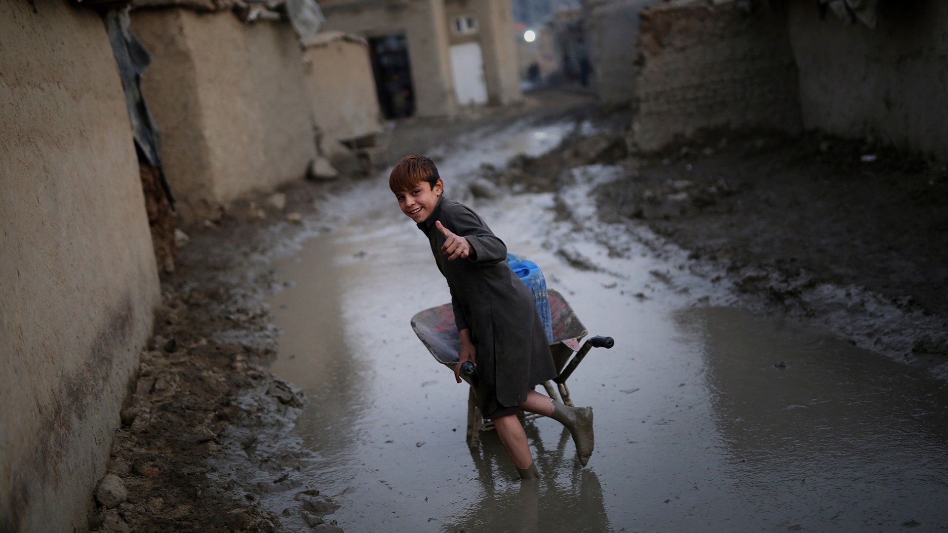 An Afghan boy gives a thumbs-up as he pushes a wheelbarrow through a muddy lane at a camp in Kabul, Afghanistan, Nov. 27, 2019.