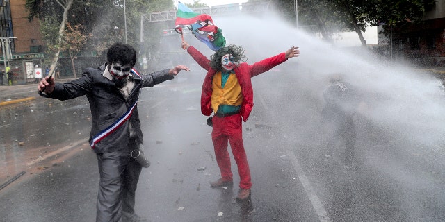 Men dressed as clowns, one dressed as the movie character “The Joker” flying a Mapuche indigenous flag, are sprayed by a police water cannon during an anti-government protest in Santiago, Chile, Monday, Nov. 4, 2019. Chile has been facing weeks of unrest, triggered by a relatively minor increase in subway fares. (AP Photo/Esteban Felix)