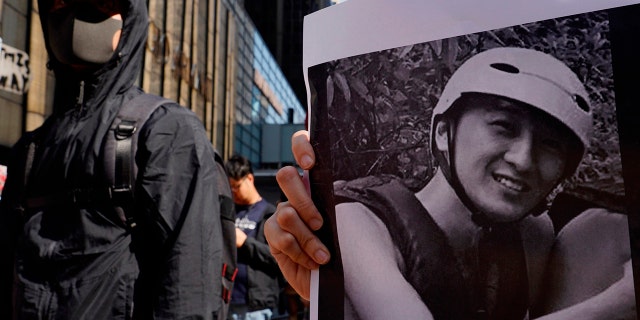 A protester stands near a photo of Chow Tsz-Lok during a memorial flash mob to remember him in Hong Kong on Friday, Nov. 8, 2019. Chow, a Hong Kong university student who fell off a parking garage after police fired tear gas during clashes with anti-government protesters died Friday, in a rare fatality after five months of unrest that intensified anger in the semi-autonomous Chinese territory. (AP Photo/Vincent Yu)