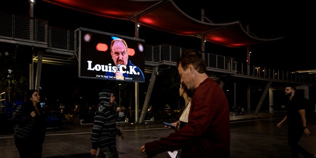 In this Thursday, Nov. 28, 2019 photo, Israelis walk past a billboard showing Comedian Louis C.K. in the Israeli city of Holon near Tel Aviv. Two years after being swept up in the Me Too movement and acknowledging sexual misconduct with multiple women, comedian Louis C.K. took to the stage at a nearly packed basketball arena outside Tel Aviv, where the audience seemed ready to let it go.(AP Photo/Tsafrir Abayov)