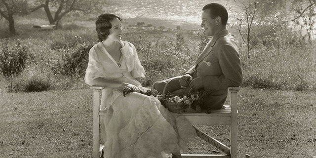 Actress Fay Wray sitting with her husband, the writer, John Monk Saunders, on a wooden bench in an outdoor setting near a lake.