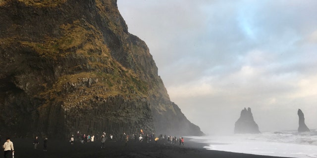 The iconic Reynisfjara Beach, as seen in October 2018, features black sand, steep cliffs, and towering Reynisdrangar sea stacks in the distance.