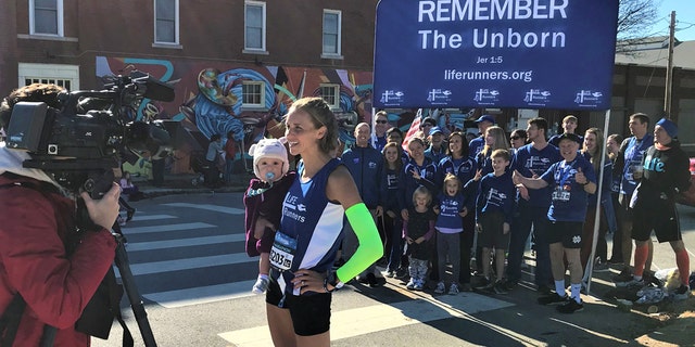 Julia Webb and her 10-month-old daughter, Gabby, talking to local media in front of her Life Runners team, a pro-life group that has members in 39 countries.