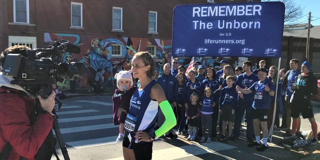 Julia Webb and her 10-month-old daughter, Gabby, talking to local media in front of her Life Runners team, a pro-life group that has members in 39 countries.