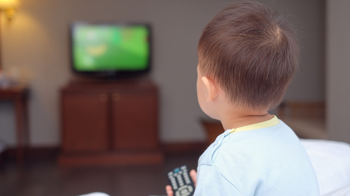 Toddler boy sitting in bed holding the tv remote control and watching television.