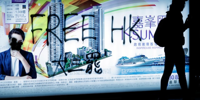 A pro-democracy protester walks by a spray-painted advertisement as demonstrators gather on a street during a protest in Hong Kong, Sunday, Dec. 8, 2019. Thousands of people took to the streets of Hong Kong on Sunday in a march seen as a test of the enduring appeal of an anti-government movement about to mark a half year of demonstrations. (AP Photo/Vincent Thian)