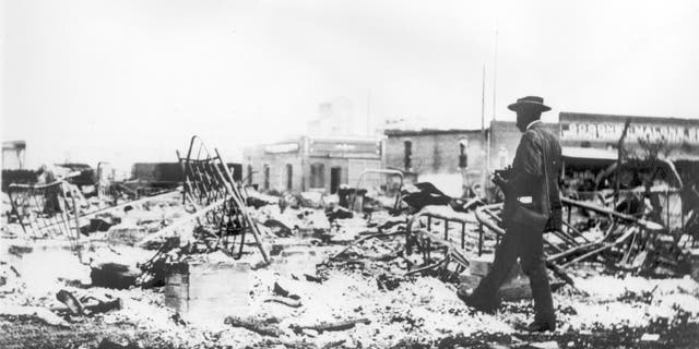 Photograph of an African American man with a camera looking at the iron bed skeletons rising above the ashes of a burnt block after the Tulsa Race Riot, Tulsa, Oklahoma, 1921.