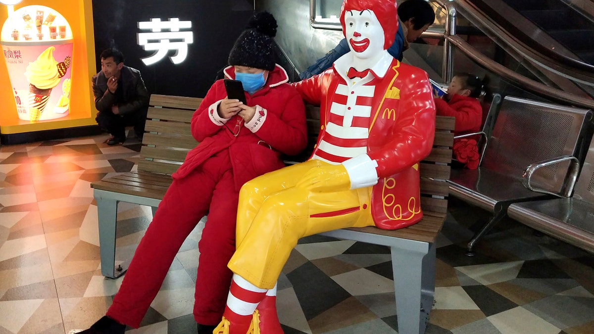 A woman wears a face mask and she uses her smartphone as she sits next to a statue of Ronald McDonald at a McDonald's restaurant in Suzhou in eastern China's Jiangsu Province on Jan. 26. (Chinatopix via AP)