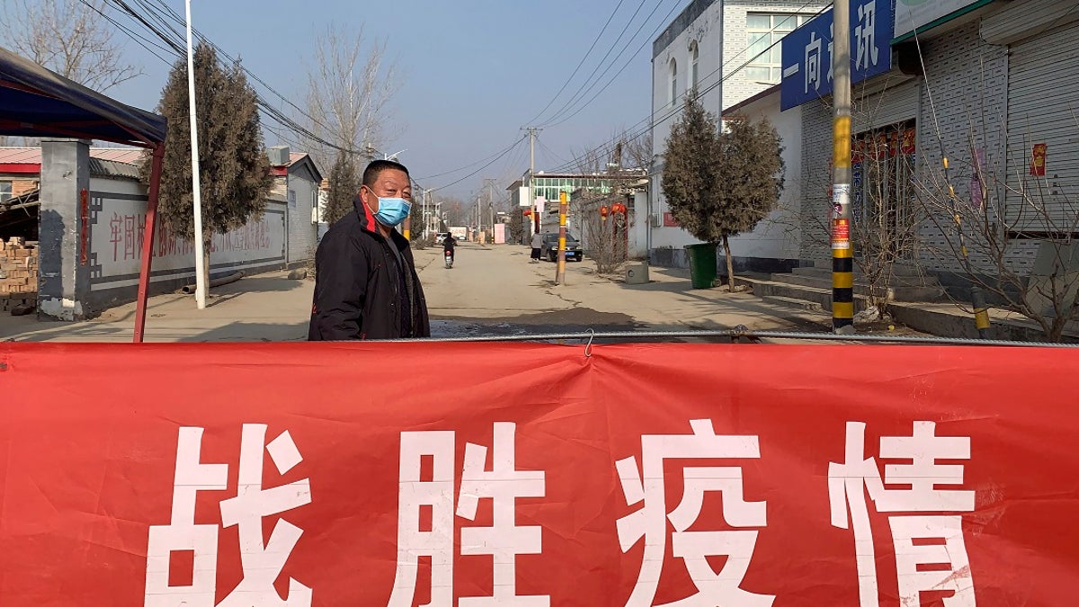 A man wearing a face mask stands near a banner reading "defeat the epidemic" stretched across the entrance to Donggouhe village in northern China's Hebei Province. (AP Photo)