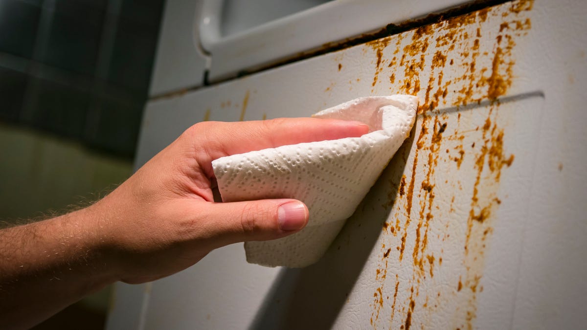 Hand cleaning baked on kitchen grime on side of oven appliance, using paper towel and cleaner.