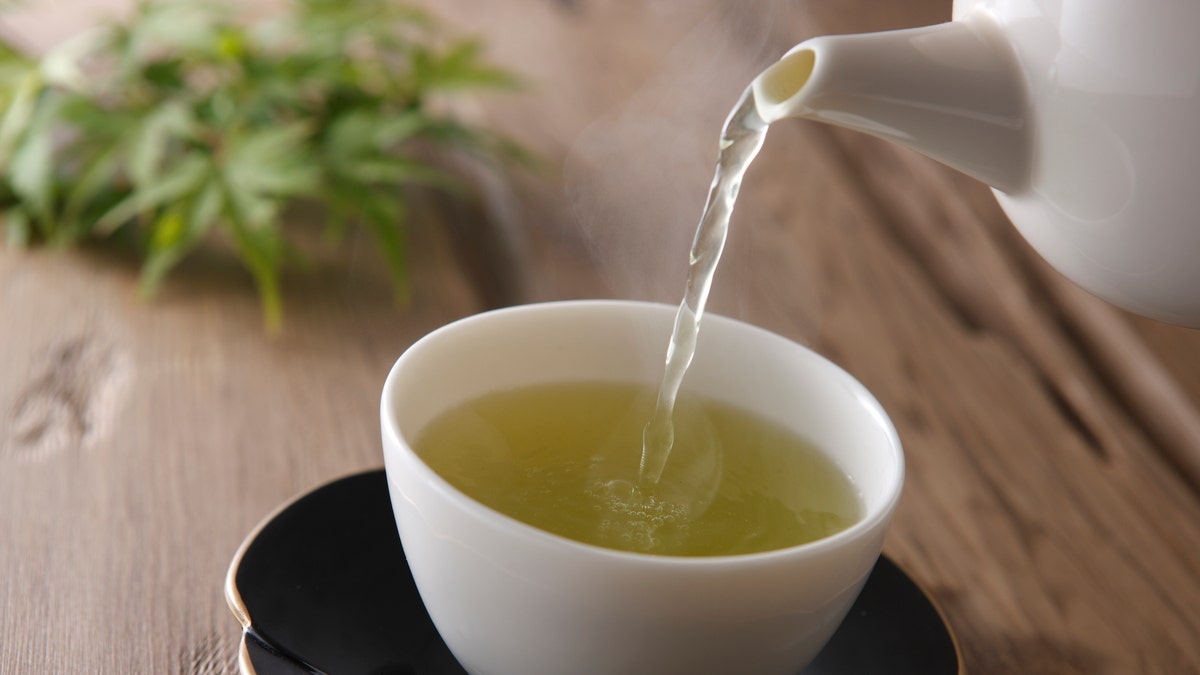 Green tea pours from spout of tea pot into porcelain mug.