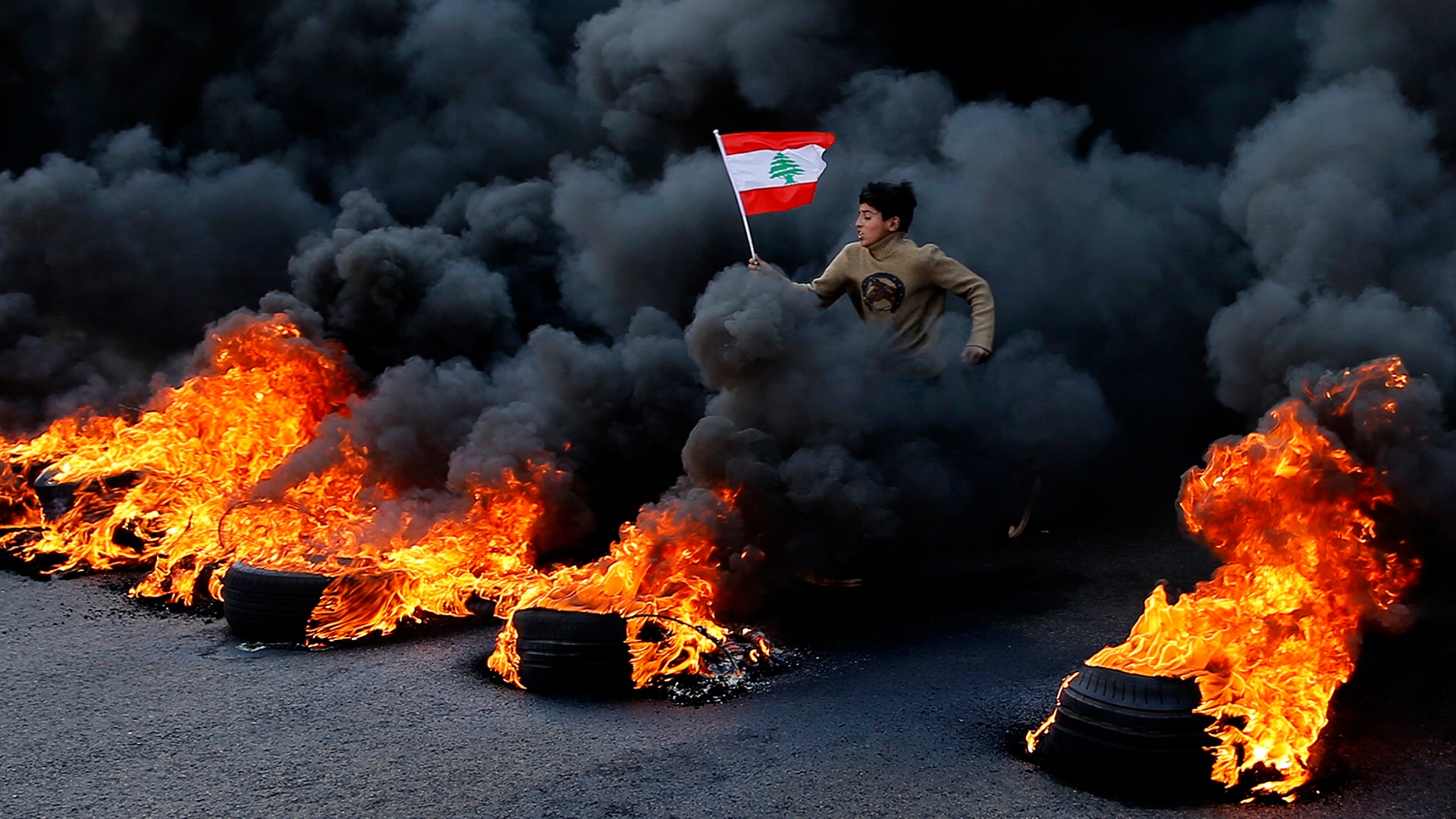 An anti-government demonstrator jumps on tires that were set on fire to block a highway as he holds a national flag, during a protest in the town of Jal el-Dib, north of Beirut, Lebanon, Jan. 14, 2020.
