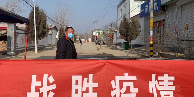 A man wearing a face mask stands near a banner reading "defeat the epidemic" stretched across the entrance to Donggouhe village in northern China's Hebei Province. (AP Photo)
