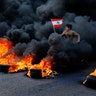 An anti-government demonstrator jumps on tires that were set on fire to block a highway as he holds a national flag, during a protest in the town of Jal el-Dib, north of Beirut, Lebanon, Jan. 14, 2020.