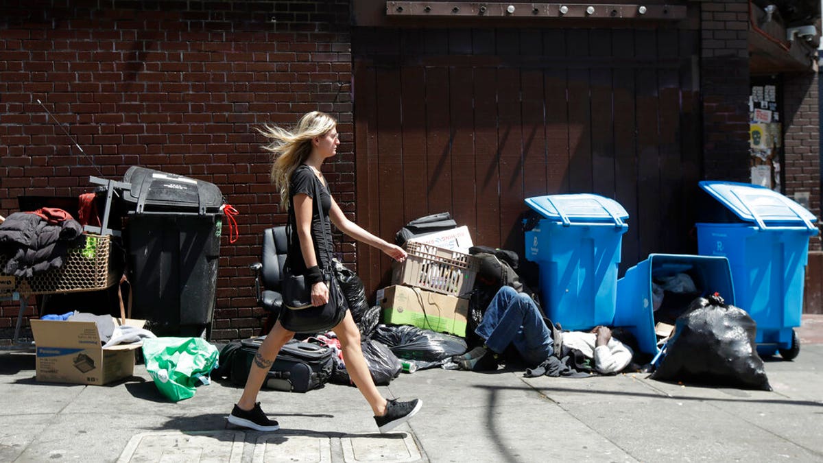 A woman walks past a homeless man sleeping in front of recycling bins and garbage on a street corner in San Francisco. 