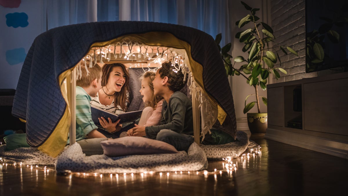Young cheerful nanny reading stories to small kids in a tent at home.