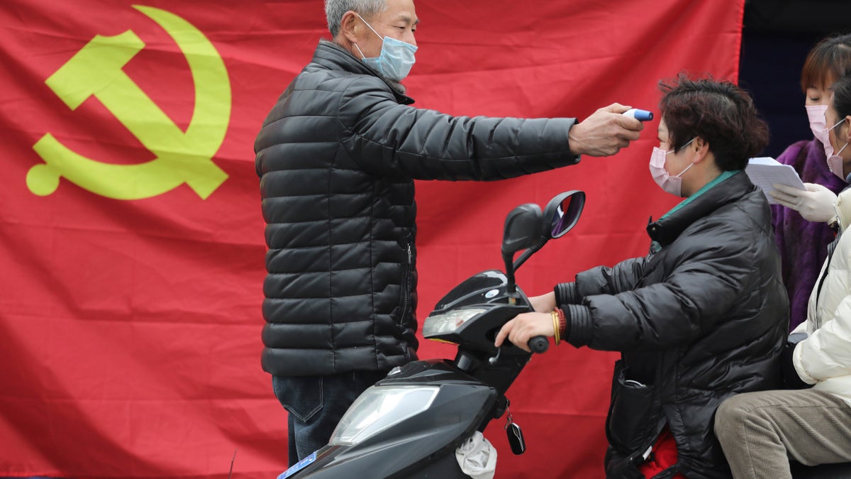 A volunteer stands in front of a Communist Party flag as he takes the temperature of a scooter driver at a roadside checkpoint in Hangzhou in eastern China's Zhejiang Province, Monday, Feb. 3, 2020. China sent medical workers and equipment to a newly built hospital, infused cash into financial markets and further restricted people's movement in sweeping new steps Monday to contain a rapidly spreading virus and its escalating impact. (Chinatopix via AP)